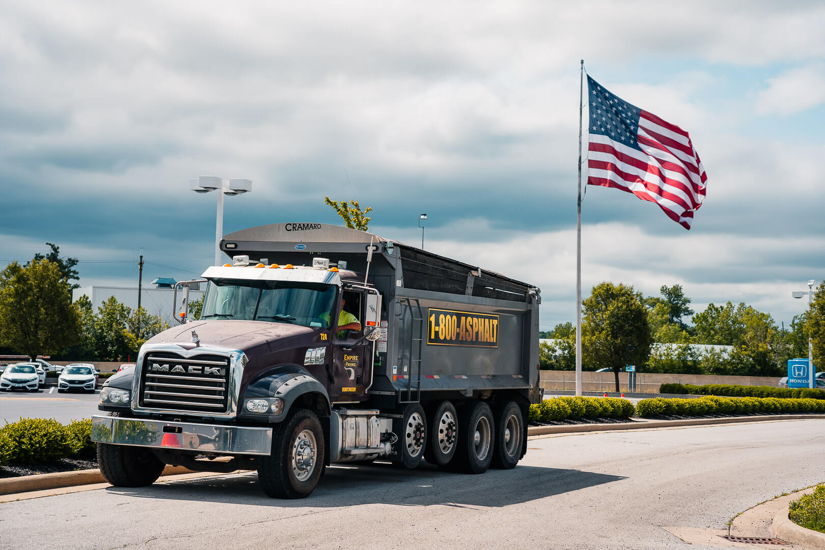 A black Mack dump truck with 1-800-ASPHALT on the side, from one of the top asphalt companies Cleveland Ohio, is parked on a road near landscaping. An American flag waves on a tall pole in the background under a partly cloudy sky.