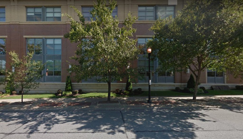 A brick building with large windows is partially obscured by leafy trees and shrubs on a sunny day. The sidewalk and street in front, expertly maintained by asphalt companies Cleveland Ohio, are shaded by tree shadows.