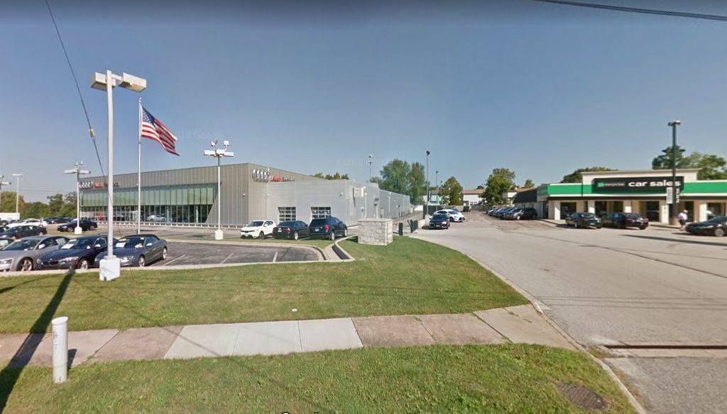 A car dealership with several parked vehicles sits beside a grey building labeled “Sissel,” a green and white car sales office, and smooth pavement—showcasing quality work from commercial asphalt companies near me—under a clear blue sky and an American flag.
