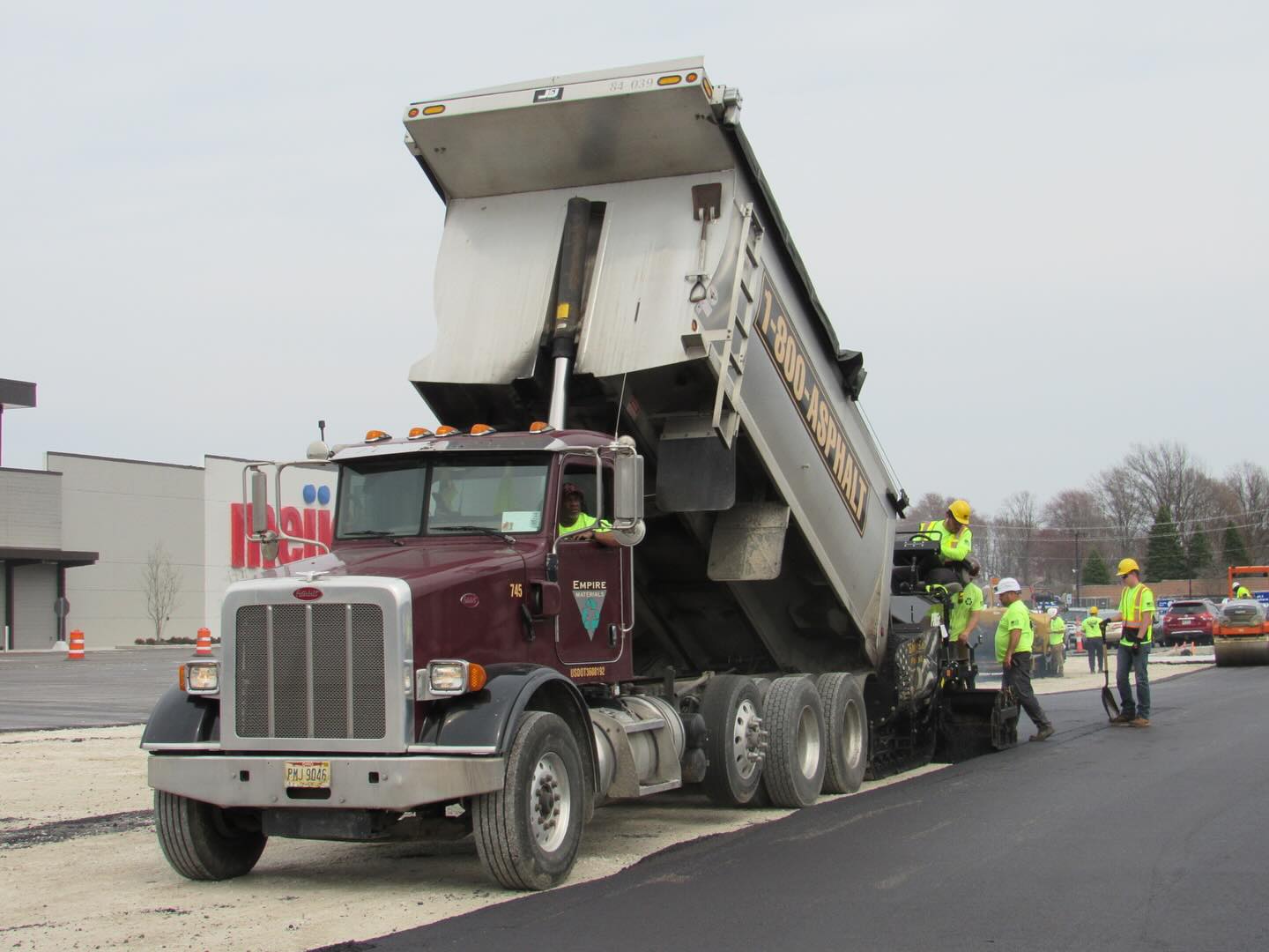 A dump truck unloads asphalt into a paving machine as several construction workers in safety gear, part of a leading commercial asphalt company near me, guide the process and work on freshly laid pavement by the roadside.
