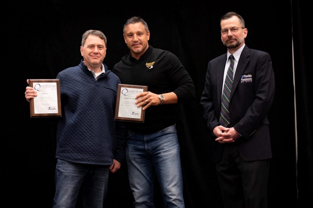 Three men pose for a photo against a black background; two hold award plaques, while the third, dressed in a suit and tie, stands with hands clasped in front of him.