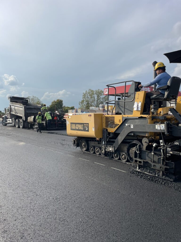 Construction workers in safety gear are operating heavy machinery to lay fresh asphalt on a road. A dump truck is unloading material, while a paver spreads it. Trees and a clear sky are visible in the background.
