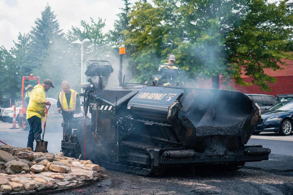Workers in yellow safety vests operate a large asphalt paving machine emitting steam, resurfacing a road near parked cars and trees on a cloudy day.