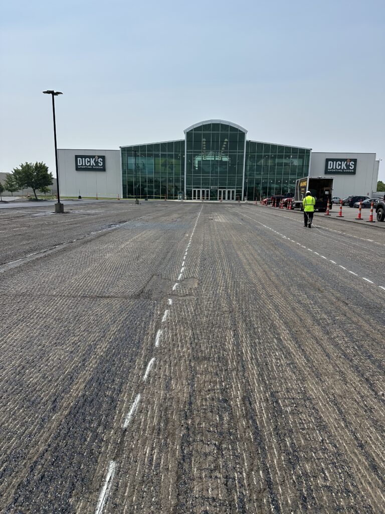 A large Dicks Sporting Goods store with a glass entrance is seen in the background. In the foreground, a rough, partially repaved parking lot stretches ahead, with a worker in a yellow vest and orange cones on the right.