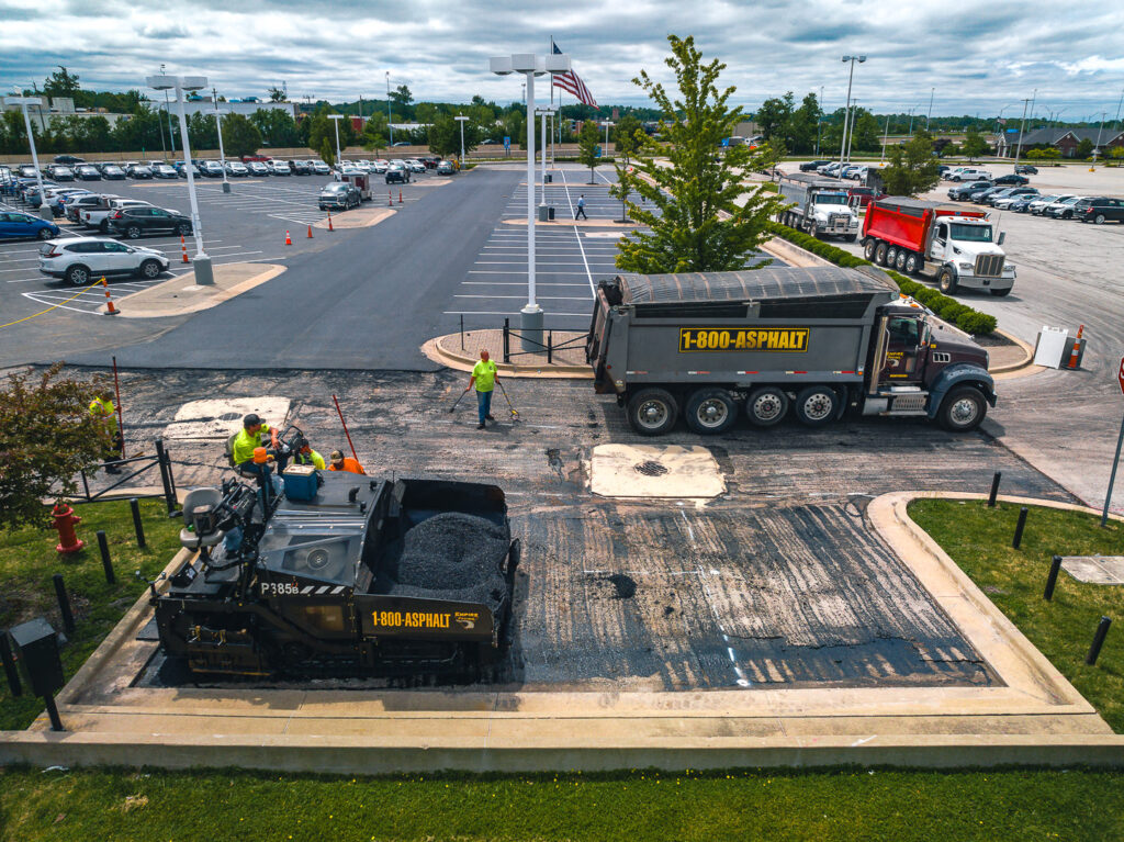 Workers repave a parking lot using heavy machinery; a dump truck labeled 1-800-ASPHALT unloads materials while several workers direct the process and guide an asphalt paver. Cars are parked in the background.