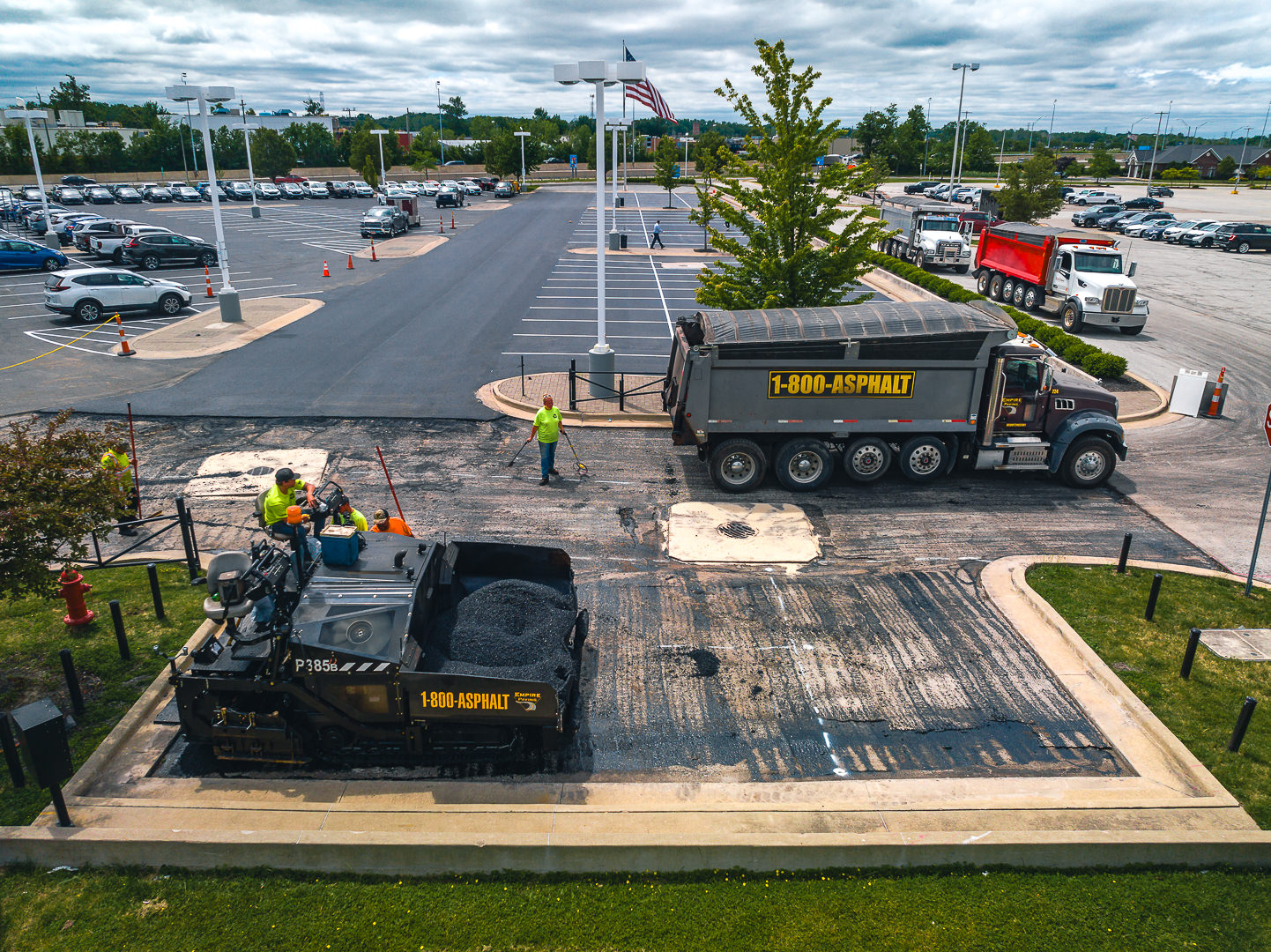 Workers repave a parking lot using heavy machinery; a dump truck labeled 1-800-ASPHALT unloads materials while several workers direct the process and guide an asphalt paver. Cars are parked in the background.