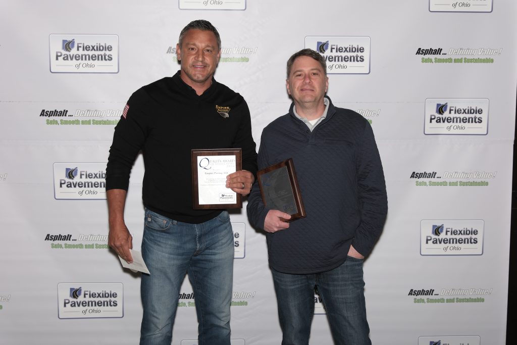Two men stand side by side, smiling and holding plaques in front of a Flexible Pavements of Ohio step-and-repeat banner. Both are dressed casually in jeans and sweaters.
