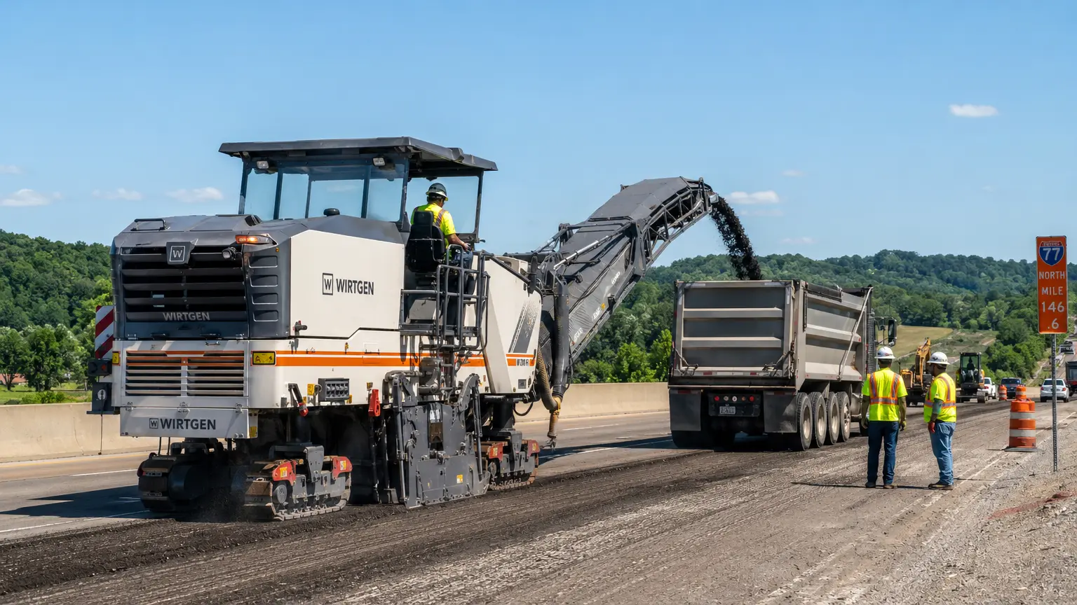 A road construction crew operates heavy machinery to mill old asphalt and load it into a dump truck on a sunny day, with workers in safety vests and traffic cones nearby.