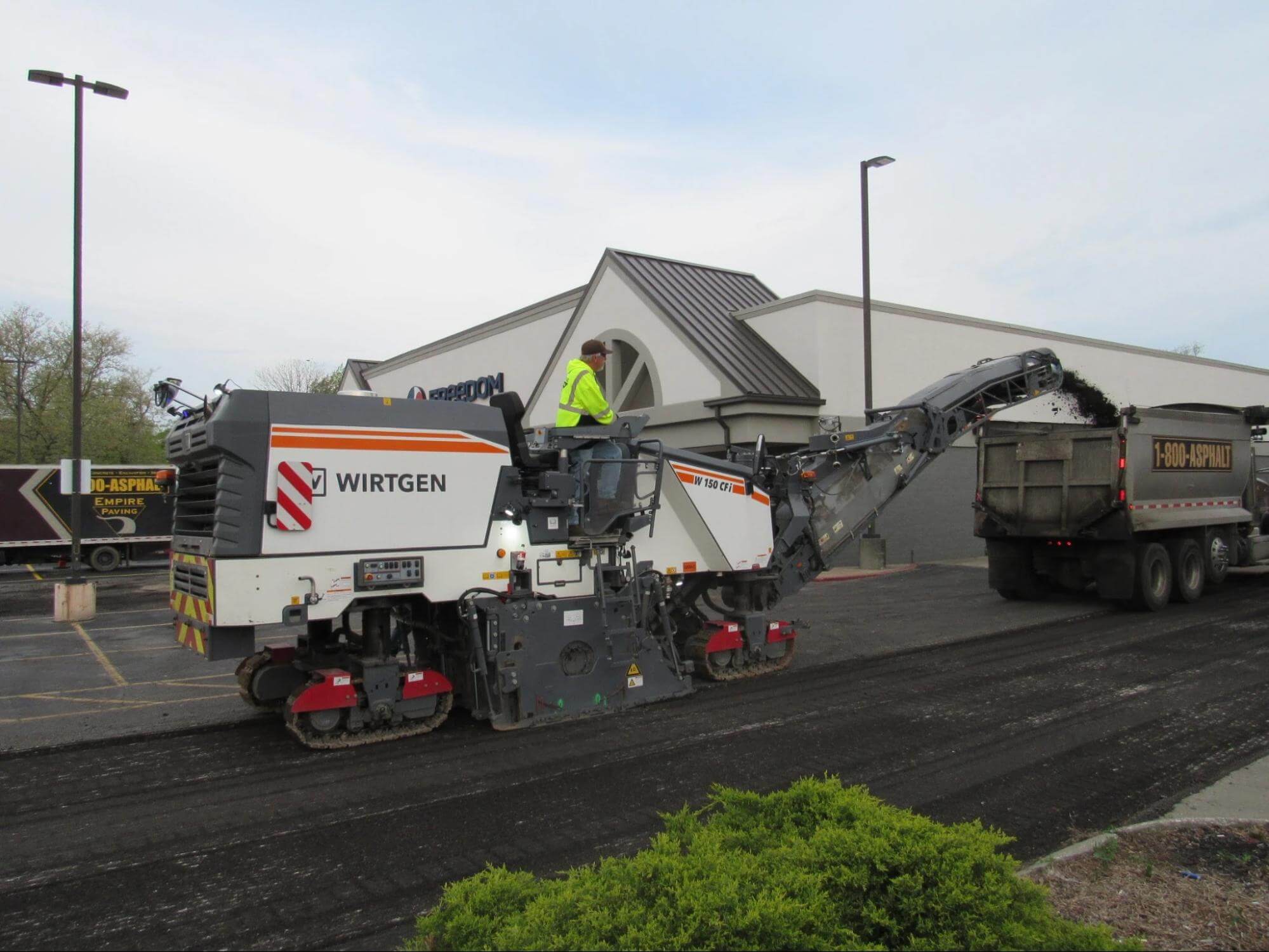 A worker operates a Wirtgen road milling machine, loading removed asphalt into a dump truck beside a commercial building in a parking lot.