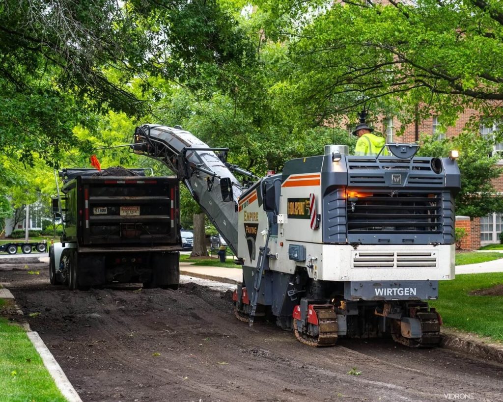 A road milling machine removes old asphalt and deposits it into a dump truck on a tree-lined residential street, with one worker in a safety vest operating the machinery.