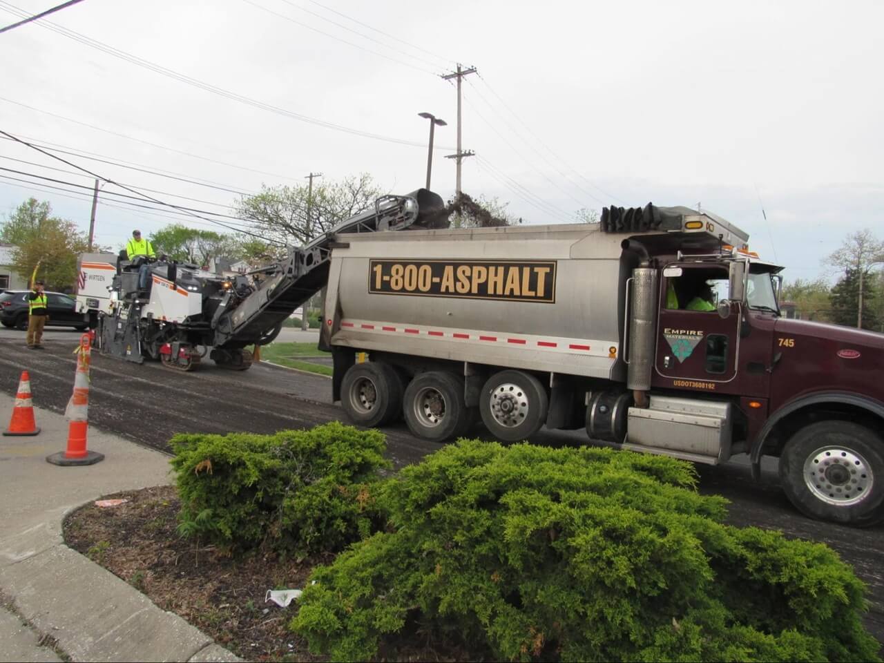 A large dump truck labeled 1-800-ASPHALT collects asphalt from a road milling machine operated by workers in safety gear. Traffic cones are placed nearby, and green bushes are in the foreground.
