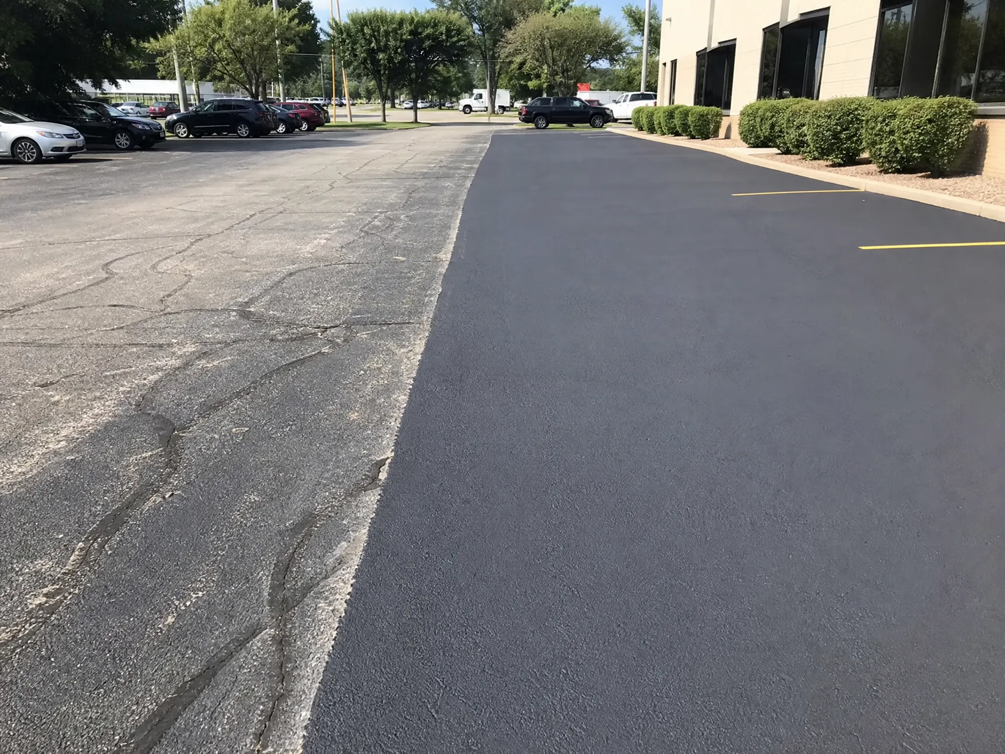 A parking lot with a clear contrast between old, cracked asphalt on the left and newly paved, smooth black asphalt on the right, next to a building with bushes.