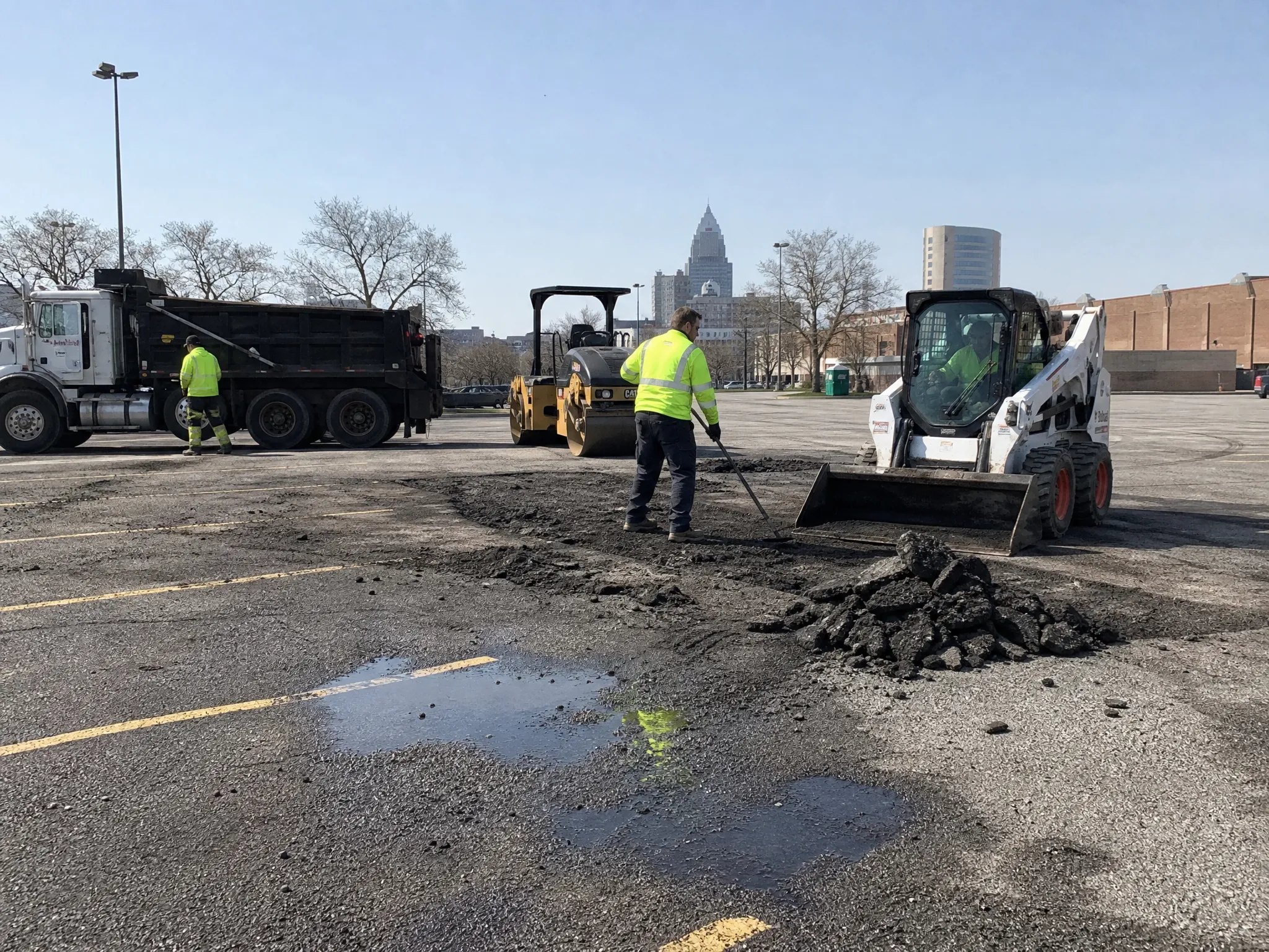 Workers in safety vests repair an asphalt parking lot using machinery and hand tools; a dump truck and roller are nearby, with a city skyline visible in the background.