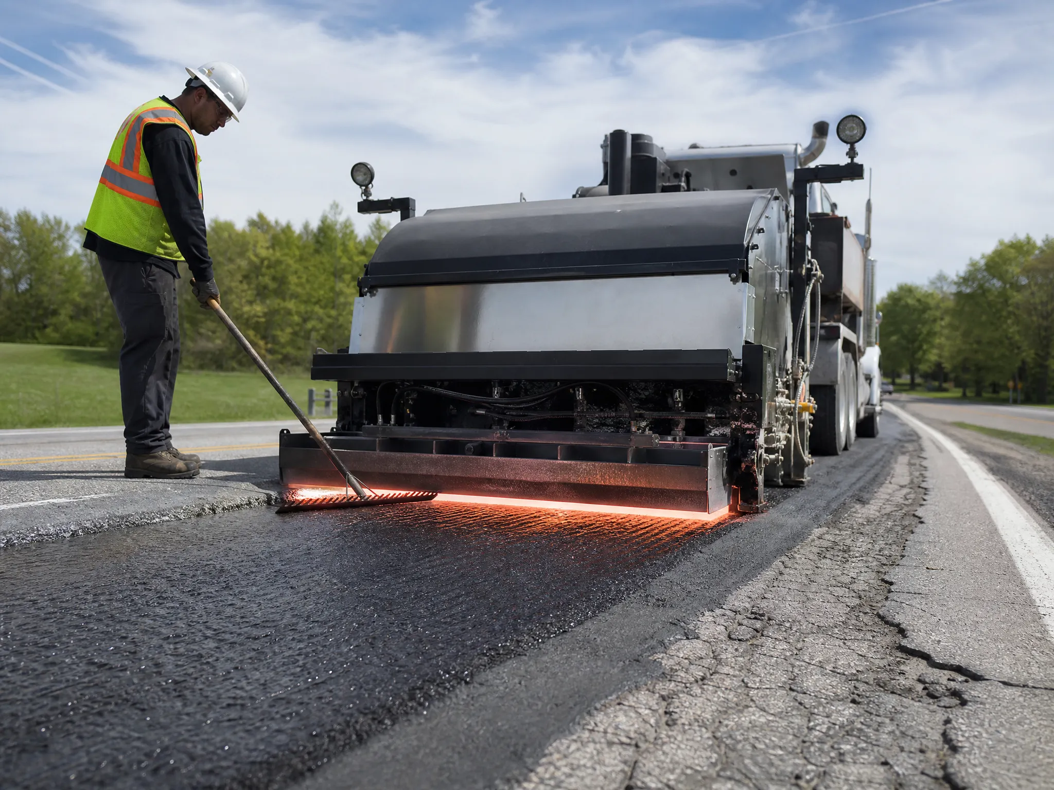 A construction worker in a safety vest and helmet uses a tool beside a large road paving machine applying hot asphalt on a rural road under a blue sky. Trees and grass are visible in the background.