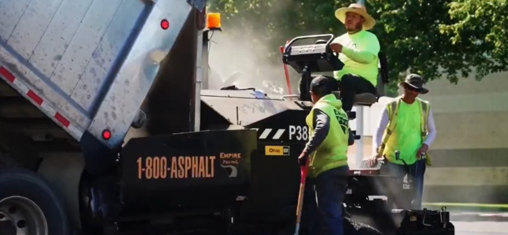 Three construction workers in safety vests operate an asphalt paving machine next to a dump truck, laying fresh asphalt on a sunny day. One worker sits on the machine, while two stand nearby, one holding a tool.