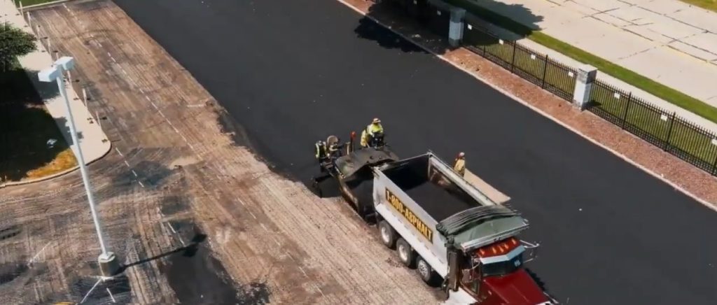 Aerial view of workers resurfacing a road, with three construction workers standing near a dump truck unloading asphalt onto the street next to a black metal fence and sidewalk.