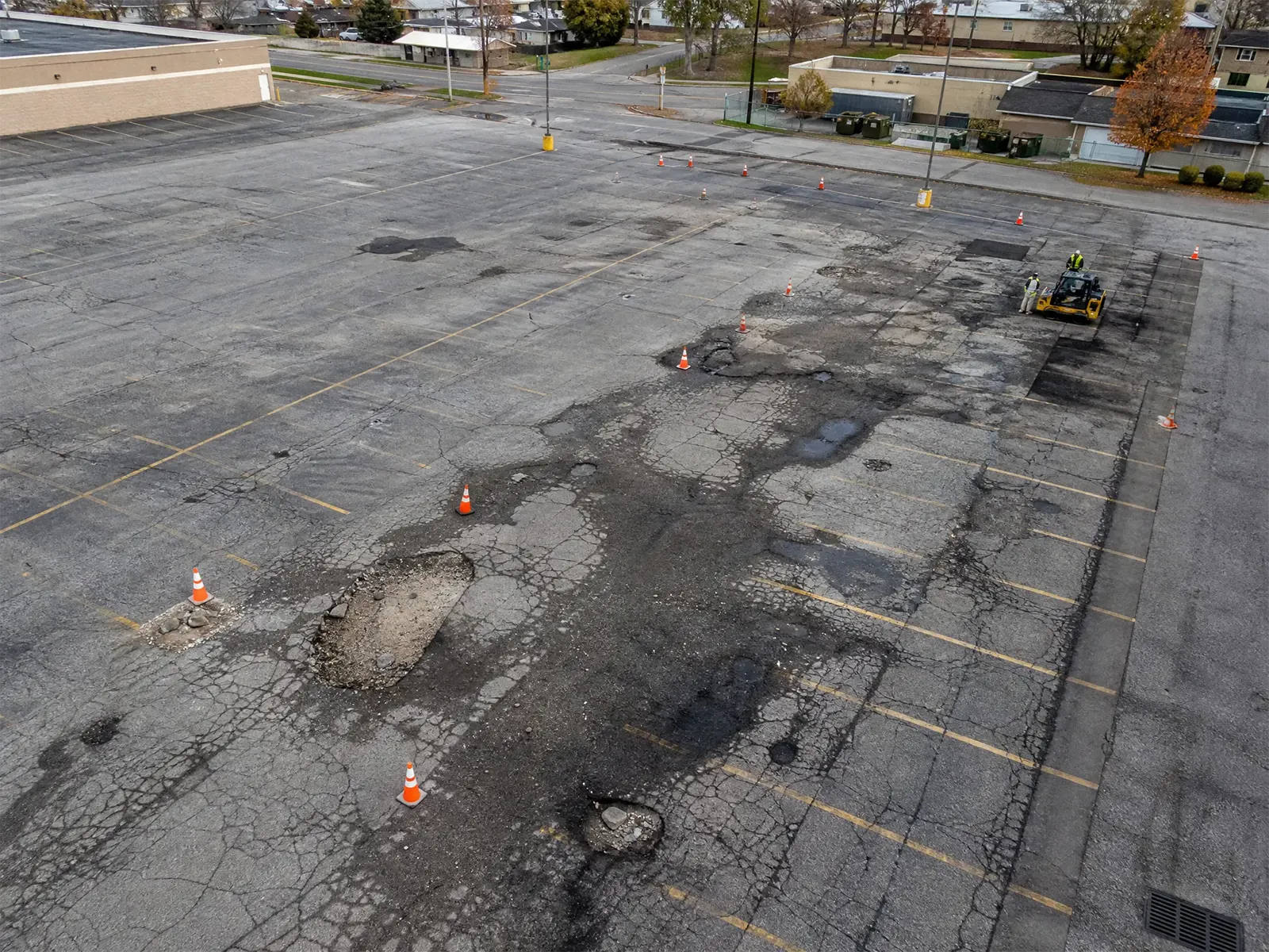 A large, mostly empty parking lot with many cracks and potholes. Orange traffic cones mark damaged areas, and a construction vehicle is working to repair part of the pavement.