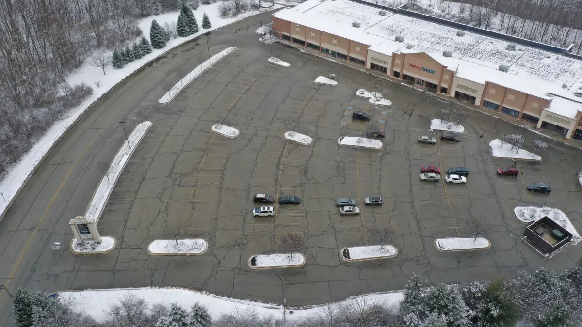 Aerial view of a mostly empty, snow-dusted parking lot in front of a large retail store. Few cars are scattered across the lot, and patches of snow line the islands and edges. Trees border the scene.