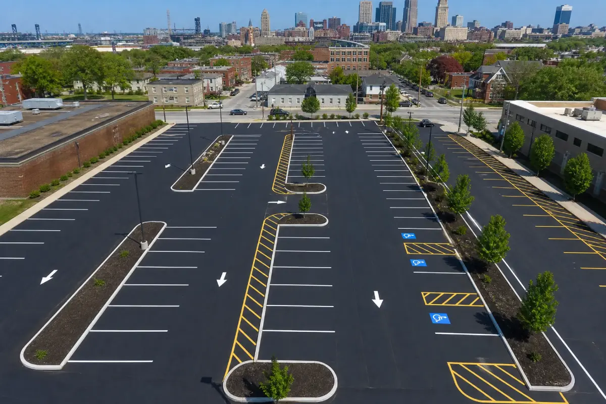 Aerial view of an empty, freshly paved parking lot with marked spaces, landscaped islands with trees, and several accessible parking spots. City buildings are visible in the background under a clear sky.