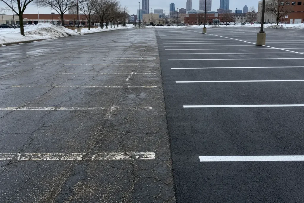 An empty parking lot shows old, cracked pavement on the left and newly paved, smooth asphalt with fresh white lines on the right. Snow piles border the lot, and a city skyline is visible in the distance.