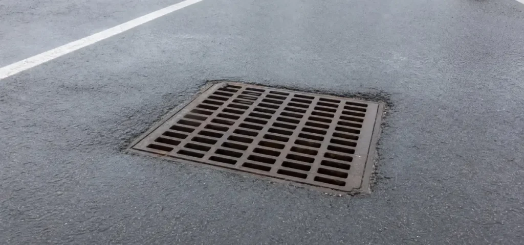 A square metal storm drain cover set into wet asphalt, with white parking lot lines visible nearby.