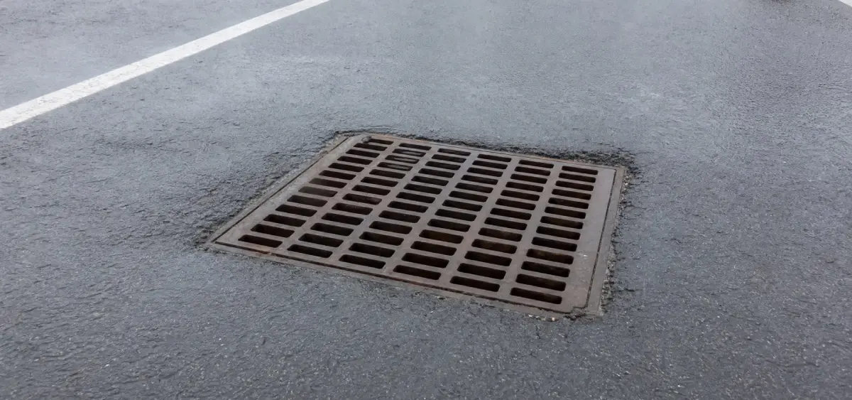 A square metal storm drain cover set into wet asphalt, with white parking lot lines visible nearby.