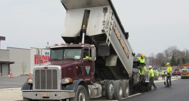 A dump truck unloads asphalt into a paving machine as several construction workers in safety gear, part of a leading commercial asphalt company near me, guide the process and work on freshly laid pavement by the roadside.
