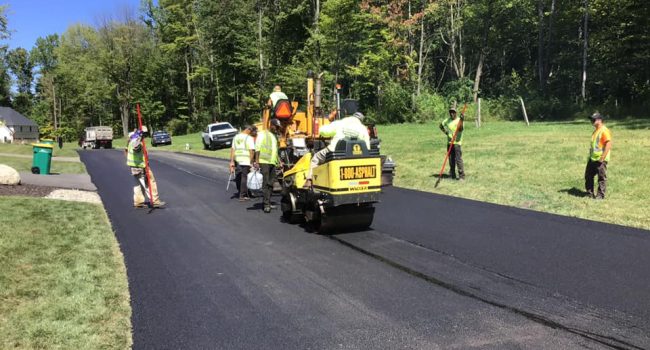 Workers in safety vests operate paving machinery for Ohio Paving and Construction, laying fresh asphalt on a rural road amid green grass and trees on a sunny day. Several workers from this commercial concrete paving contractor stand roadside.
