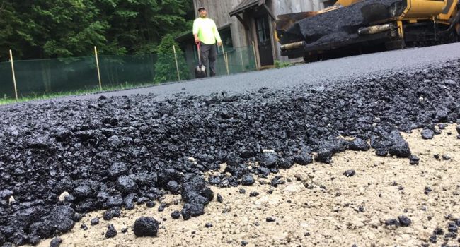 Close-up of fresh asphalt being laid over a dirt road by a yellow paving machine from Ohio Paving and Construction, with a worker in a neon yellow shirt nearby and a wooden building and trees in the background.