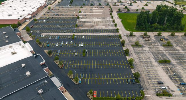 Aerial view of a large, mostly empty shopping mall parking lot with a few scattered cars, surrounded by trees and commercial buildings—expertly maintained by Ohio paving and construction professionals.