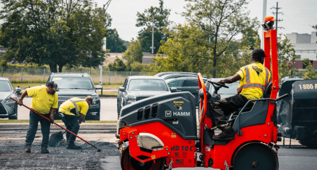 Three workers in yellow vests repave a parking lot; two use tools to spread asphalt while one operates an orange steamroller. Empire Paving, a trusted commercial asphalt company near me, handles the project as cars and trees are visible in the background.