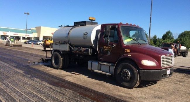 A maroon and silver tanker truck from Empire Paving applies liquid to a parking lot during daylight, likely for paving or sealing, with a shopping center and several vehicles in the background.