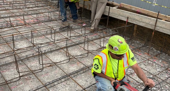 Construction workers in safety vests and helmets work on a rebar framework at a building site. One worker uses a power tool on the ground while others measure or inspect near a wooden form. Safety cones and tape are visible.