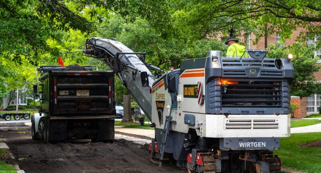 A road construction machine removes old asphalt from a street and loads it into a dump truck, while a worker in a neon vest oversees the process. Trees and residential buildings are visible in the background.