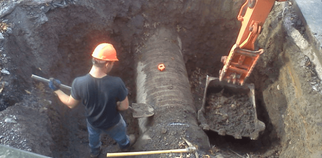 A construction worker in a hard hat stands in a deep hole beside a large, partially buried pipe while an orange excavator scoops dirt nearby, showcasing the expertise often found with commercial asphalt companies near me like Empire Paving.