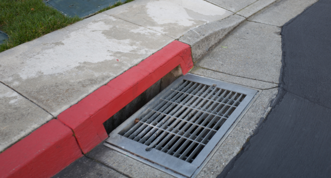 A metal storm drain grate is set into a concrete curb along a street corner, partly painted red to indicate no parking. Nearby, grass and a sidewalk hint at recent work by an experienced commercial concrete paving contractor.