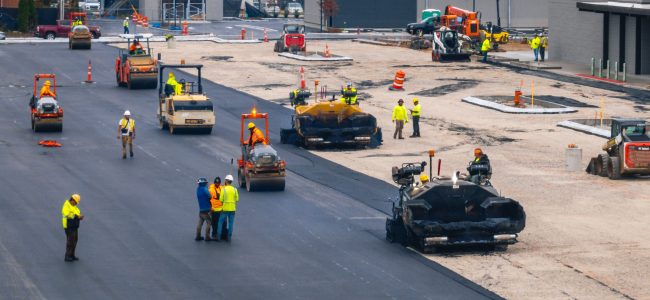 Construction workers and machinery are paving a large parking lot next to commercial buildings. Workers in safety gear operate rollers and pavers, while others discuss plans. Orange cones and barriers mark the work area.